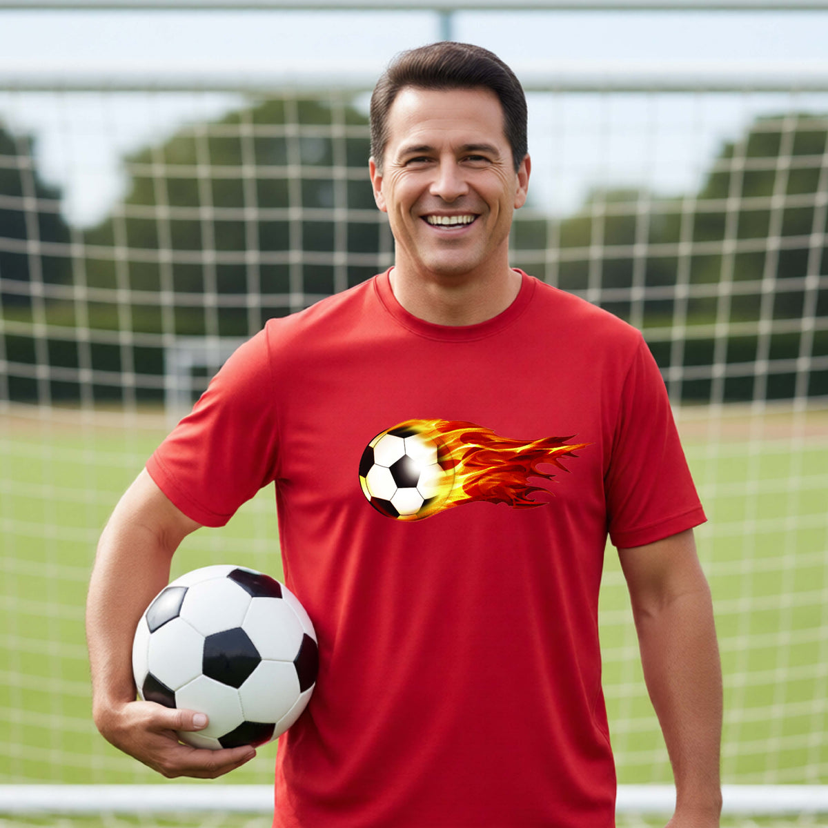A smiling man wearing a red short-sleeve t-shirt standing on a soccer field in front of a goal net. with a white graphic of a soccer ball shooting through air and trailing yellow, red and orange flames behind it, printed on the chest.