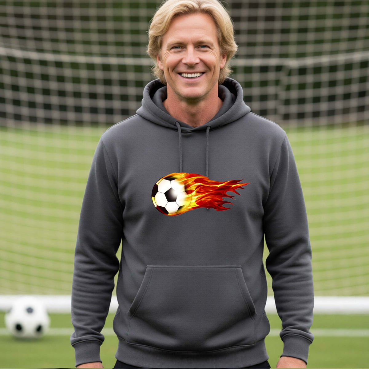 A smiling man wearing a dark gray hoodie pullover standing on a soccer field in front of a goal net. with a white graphic of a soccer ball shooting through air and trailing yellow, red and orange flames behind it, printed on the chest.