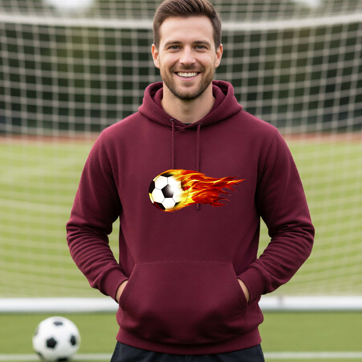 A smiling man wearing a maroon hoodie pullover standing on a soccer field in front of a goal net. with a white graphic of a soccer ball shooting through air and trailing yellow, red and orange flames behind it, printed on the chest.