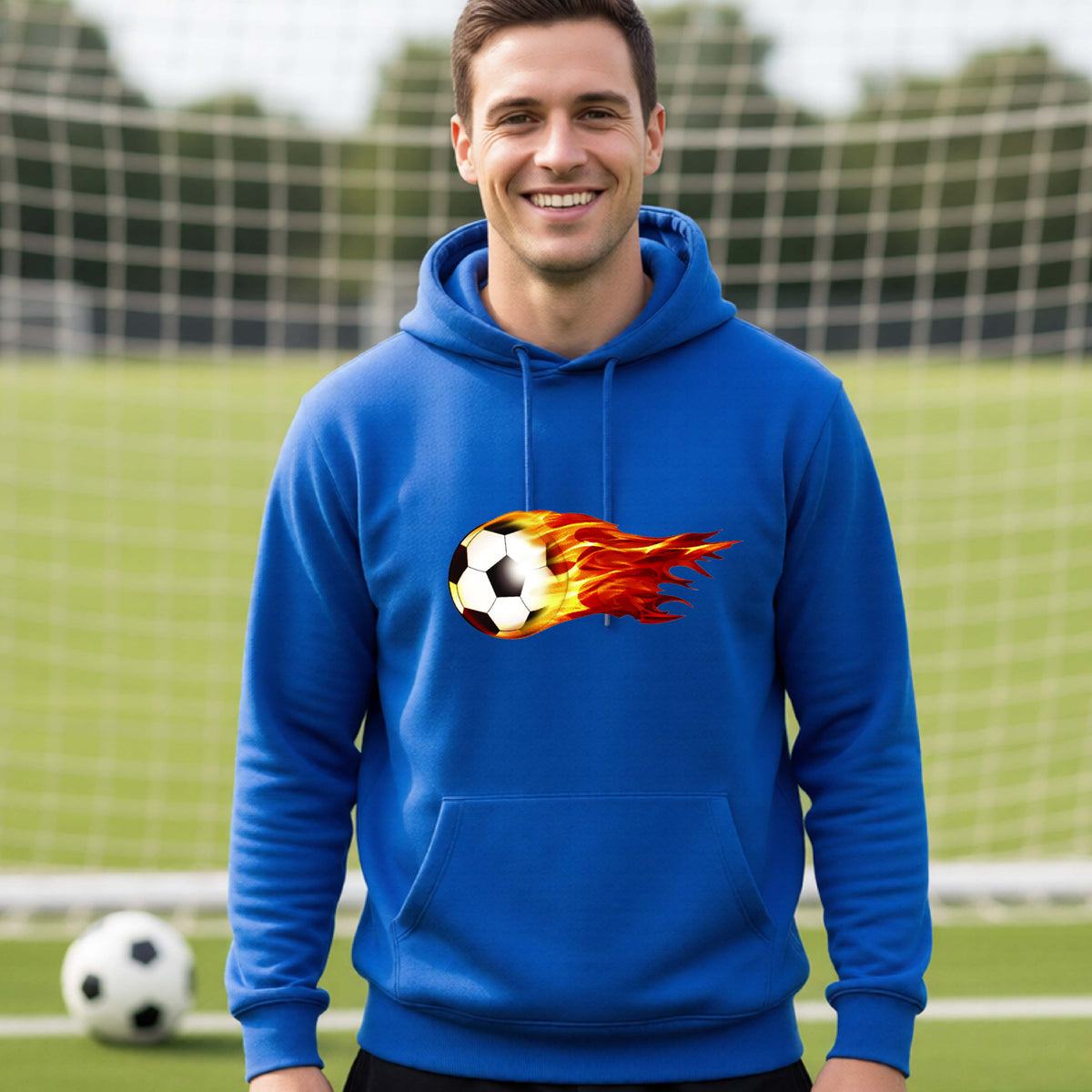 A smiling man wearing a royal blue hoodie pullover standing on a soccer field in front of a goal net. with a white graphic of a soccer ball shooting through air and trailing yellow, red and orange flames behind it, printed on the chest.