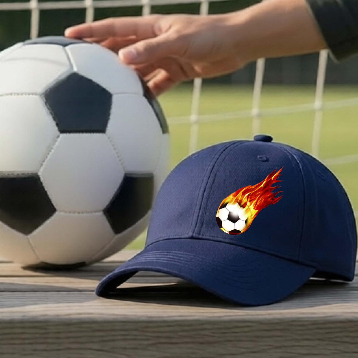 A close-up of a navy blue hat sitting on an outdoor bench next to a soccer ball with a white graphic of a soccer ball shooting through air and trailing yellow, red and orange flames behind it, printed on the front.