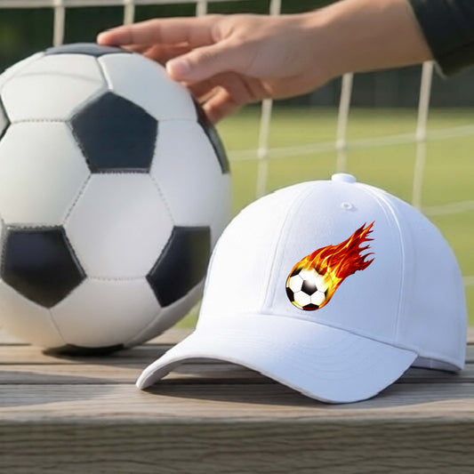 A white baseball cap with a graphic of a soccer ball engulfed in flames, displayed next to a real soccer ball on a wooden table with a hand resting on the soccer ball, set against an outdoor soccer field background.
