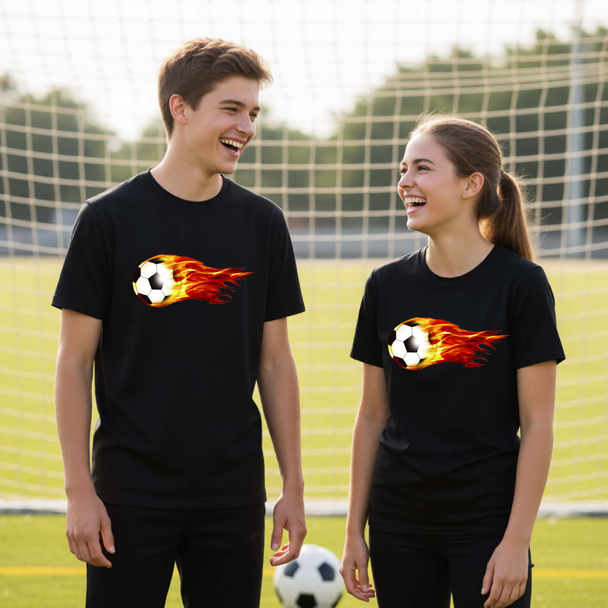 Two young athletes wearing matching black short-sleeve t-shirts standing on a soccer field in front of a goal net. with a white graphic of a soccer ball shooting through air and trailing yellow, red and orange flames behind it, printed on the chest.