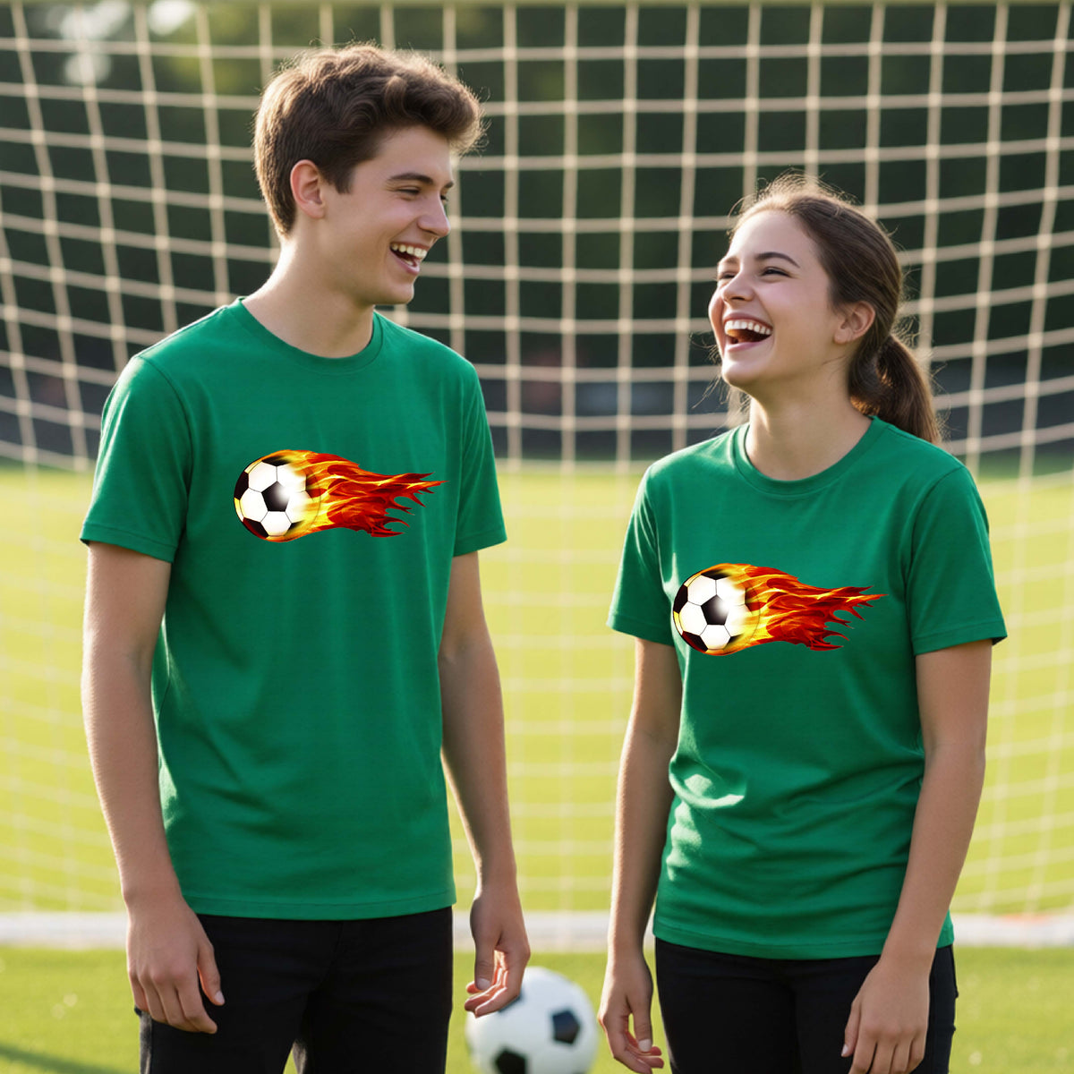 Two young athletes wearing matching green short-sleeve t-shirts standing on a soccer field in front of a goal net. with a white graphic of a soccer ball shooting through air and trailing yellow, red and orange flames behind it, printed on the chest.