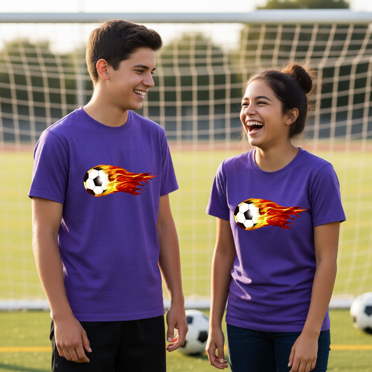 Two young athletes wearing matching purple short-sleeve t-shirts standing on a soccer field in front of a goal net. with a white graphic of a soccer ball shooting through air and trailing yellow, red and orange flames behind it, printed on the chest.
