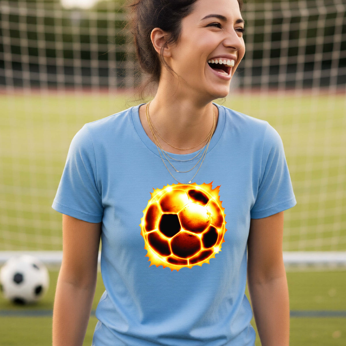 A woman laughing on a soccer field wearing a light blue t-shirt with a glowing soccer ball graphic on the chest.