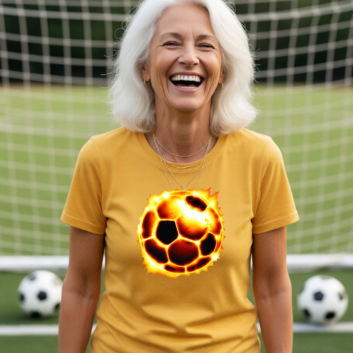 A smiling older woman with white hair stands on a soccer field in front of a goal net, wearing a mustard yellow T-shirt with a large glowing soccer ball graphic on the chest. Soccer balls are visible on the field in the background.