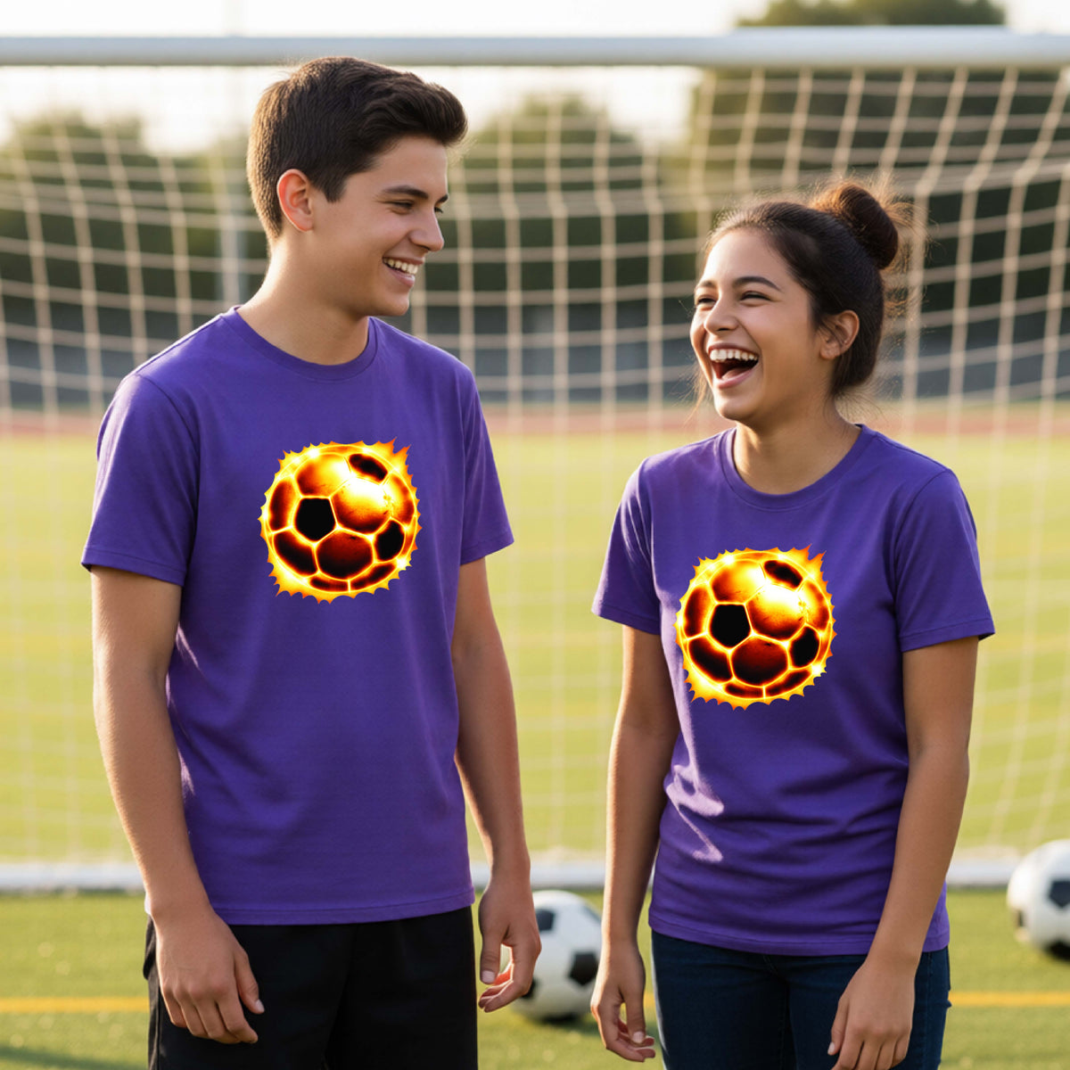 A young man and a young woman standing on a soccer field wearing matching purple t-shirts with a graphic of a soccer ball engulfed in flames. The background shows a blurred soccer goal net and grass.