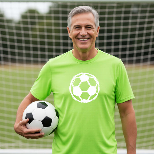 A smiling older man wearing a bright lime green t-shirt with a white soccer ball graphic on the chest, holding a black and white soccer ball on an outdoor soccer field in front of a goal net.