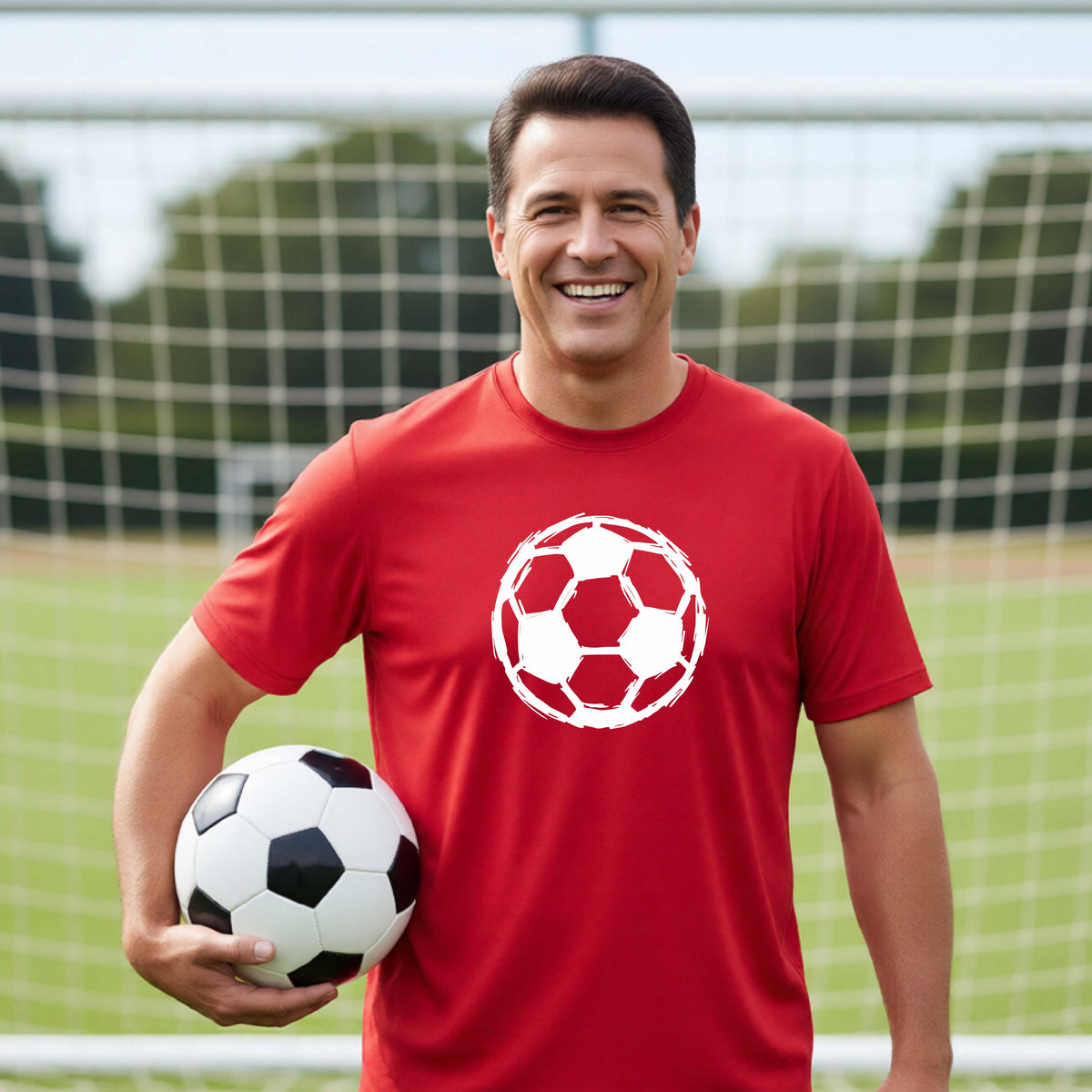 A smiling man wearing a red short-sleeve athletic T-shirt with a white soccer ball graphic on the chest, holding a black-and-white soccer ball on a grassy field in front of a goal net.