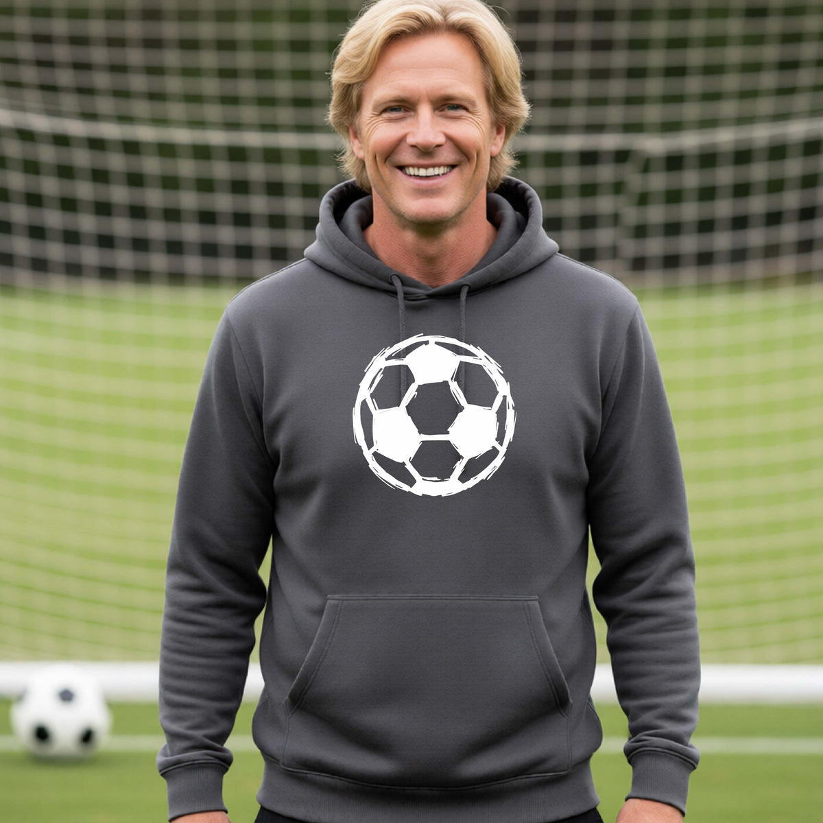 A smiling man wearing a dark gray hoodie with a white soccer ball graphic on the chest, standing on a soccer field with a goal net in the background.