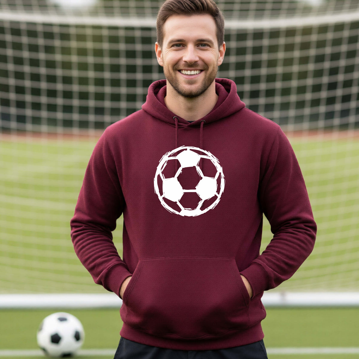 A smiling man wearing a maroon hoodie with a white soccer ball graphic on the chest, standing on a soccer field with a goal net in the background.