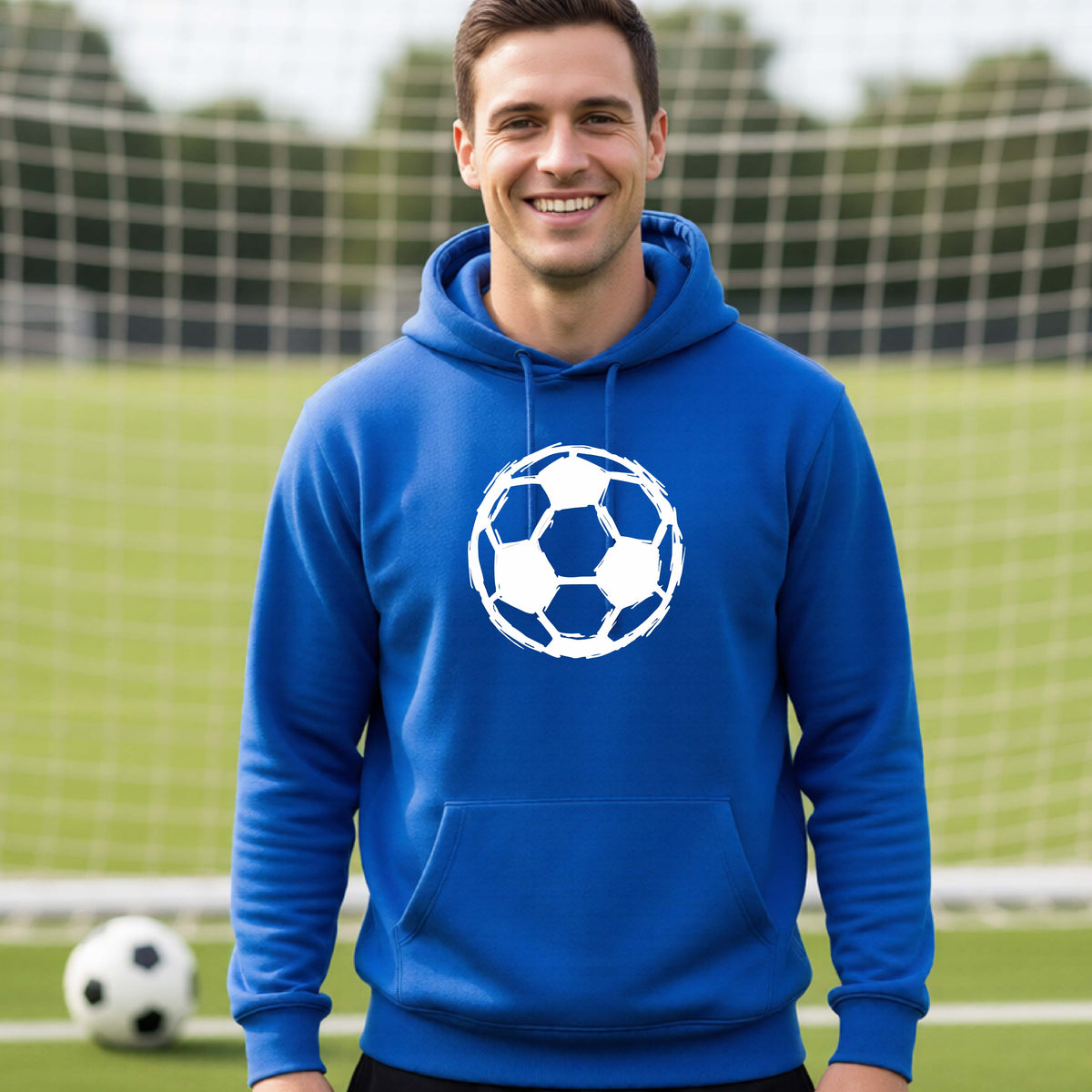 A smiling man wearing a royal blue hoodie with a white soccer ball srtistic graphic on the chest, standing on a soccer field with a goal net and ball in the background.
