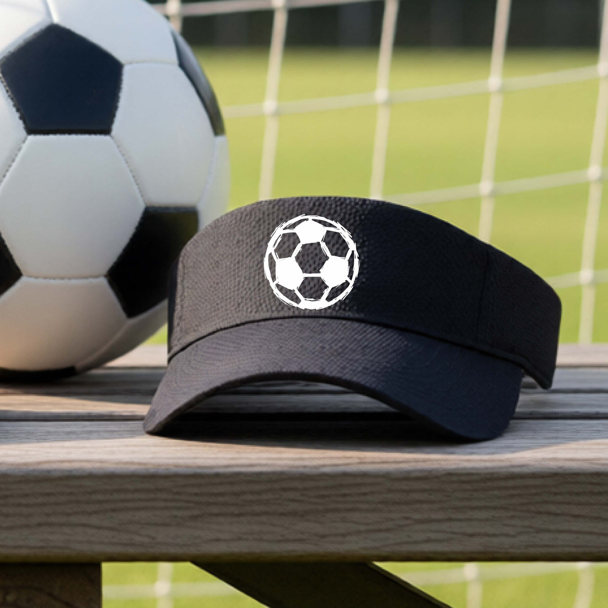 A black visor with a white soccer ball logo on the front, resting on a wooden bench with a soccer ball and a goal net in the background.
