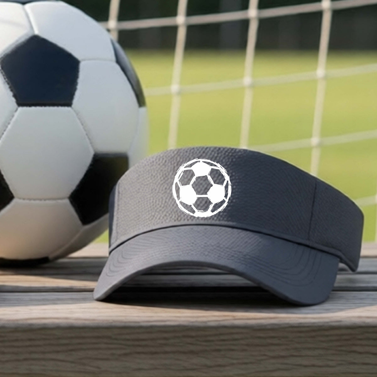 A dark gray visor with a white soccer ball graphic on the front, resting on a wooden surface with a soccer ball and goal net in the background.