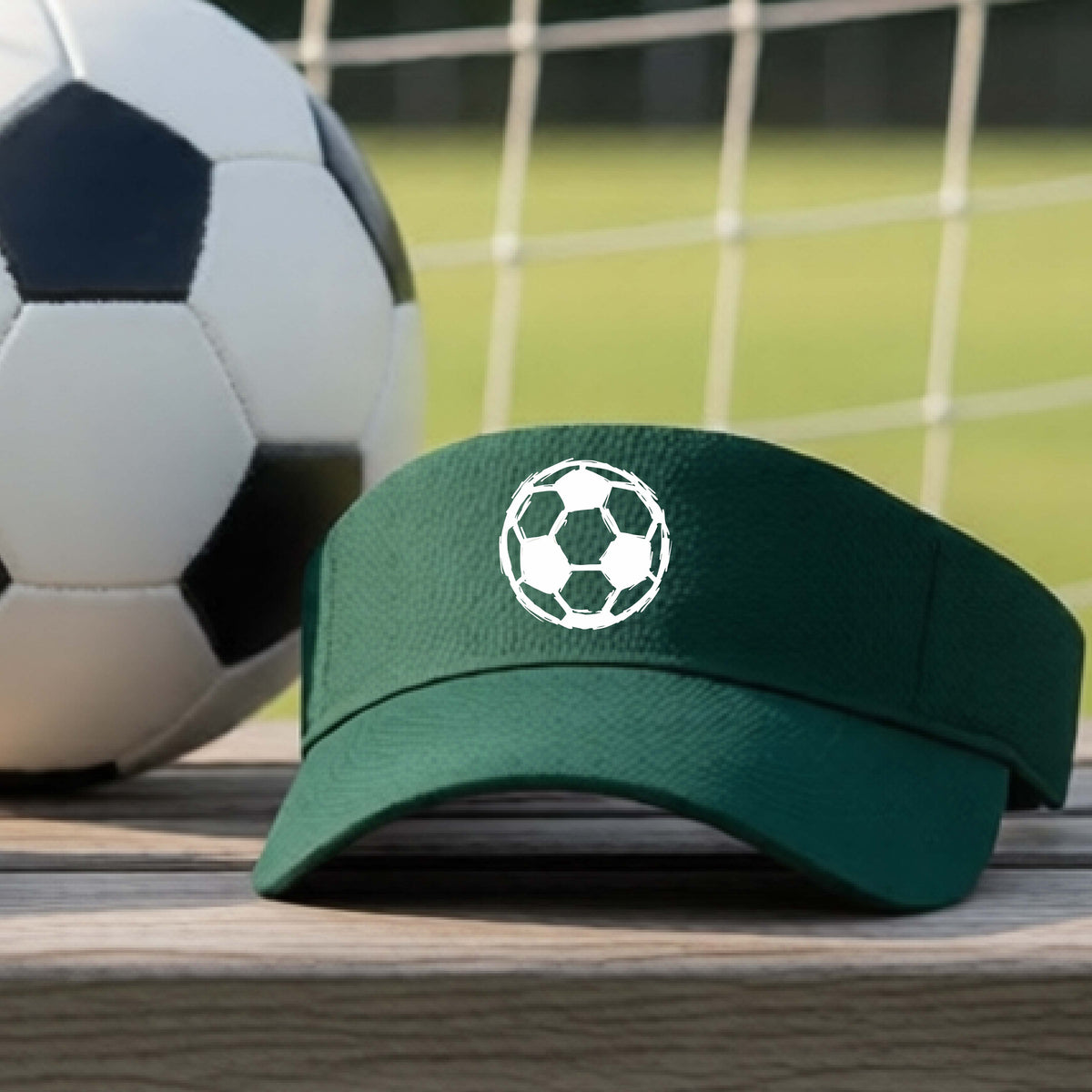 A dark green visor with a white soccer ball graphic on the front, resting on a wooden surface with a soccer ball and goal net in the background.