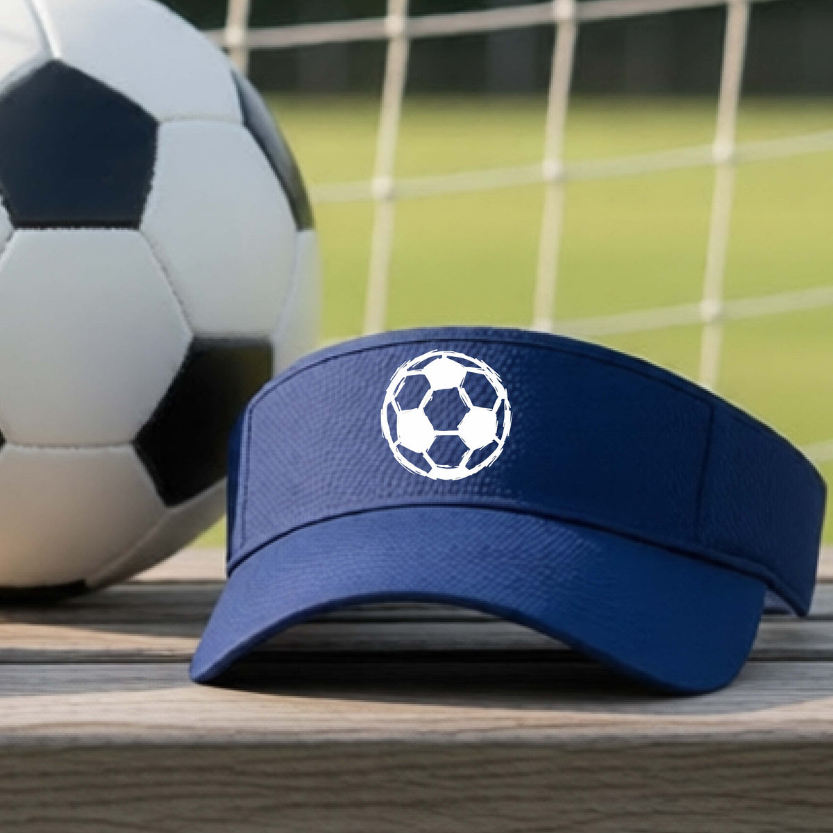 A navy blue visor with a white soccer ball graphic on the front, resting on a wooden surface with a soccer ball and a goal net in the background.