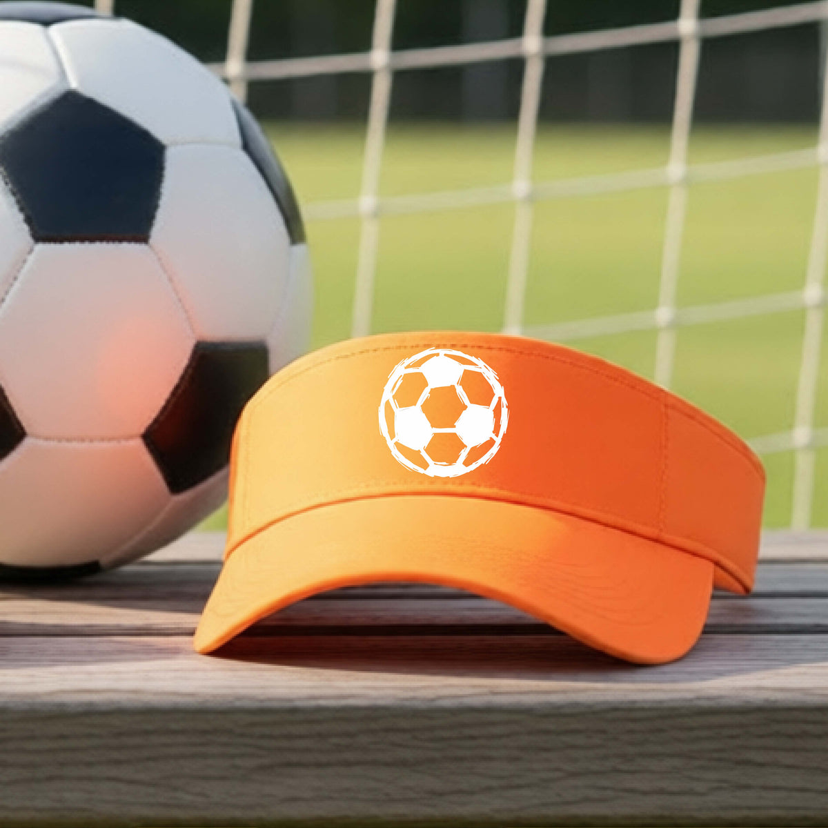 An orange visor with a white soccer ball graphic on the front, resting on a wooden surface with a soccer ball and a goal net in the background.