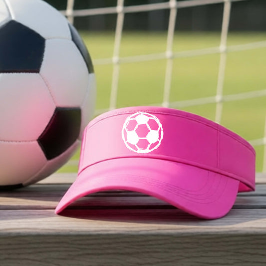 A bright pink athletic visor with a white soccer ball graphic on the front, resting on a wooden surface with a soccer ball and goal net blurred in the background.