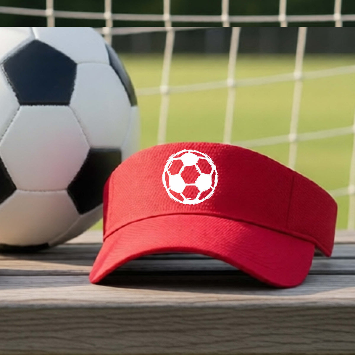 A red visor hat with a white soccer ball graphic on the front, resting on a wooden surface with a soccer ball and a goal net in the blurred background.