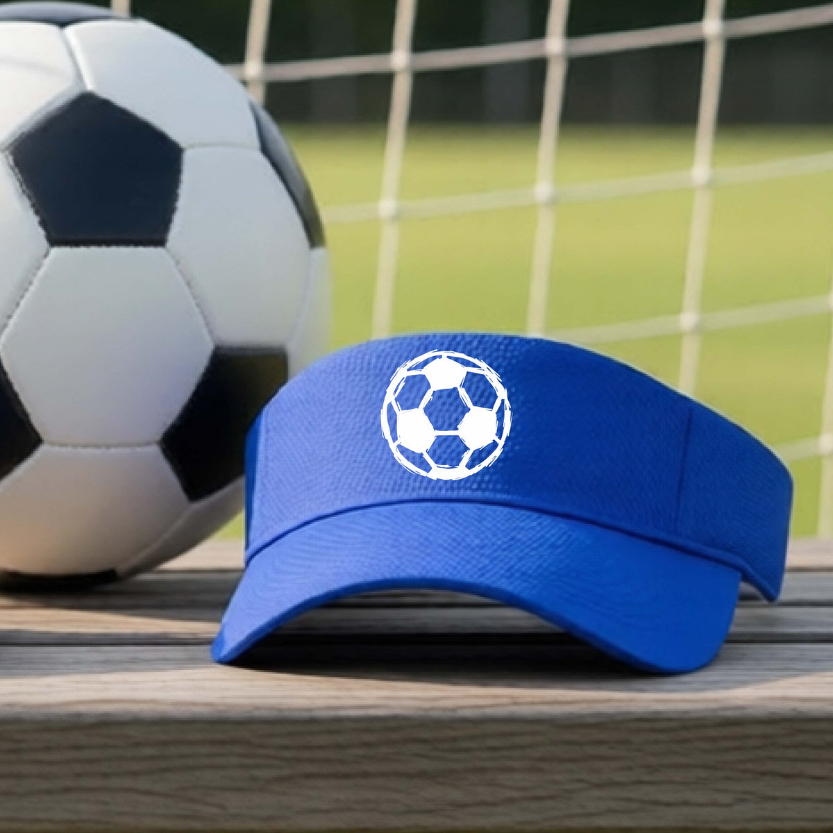 A royal blue visor with a white soccer ball graphic on the front, resting on a wooden surface next to a black and white soccer ball with a blurred soccer net and green field in the background.