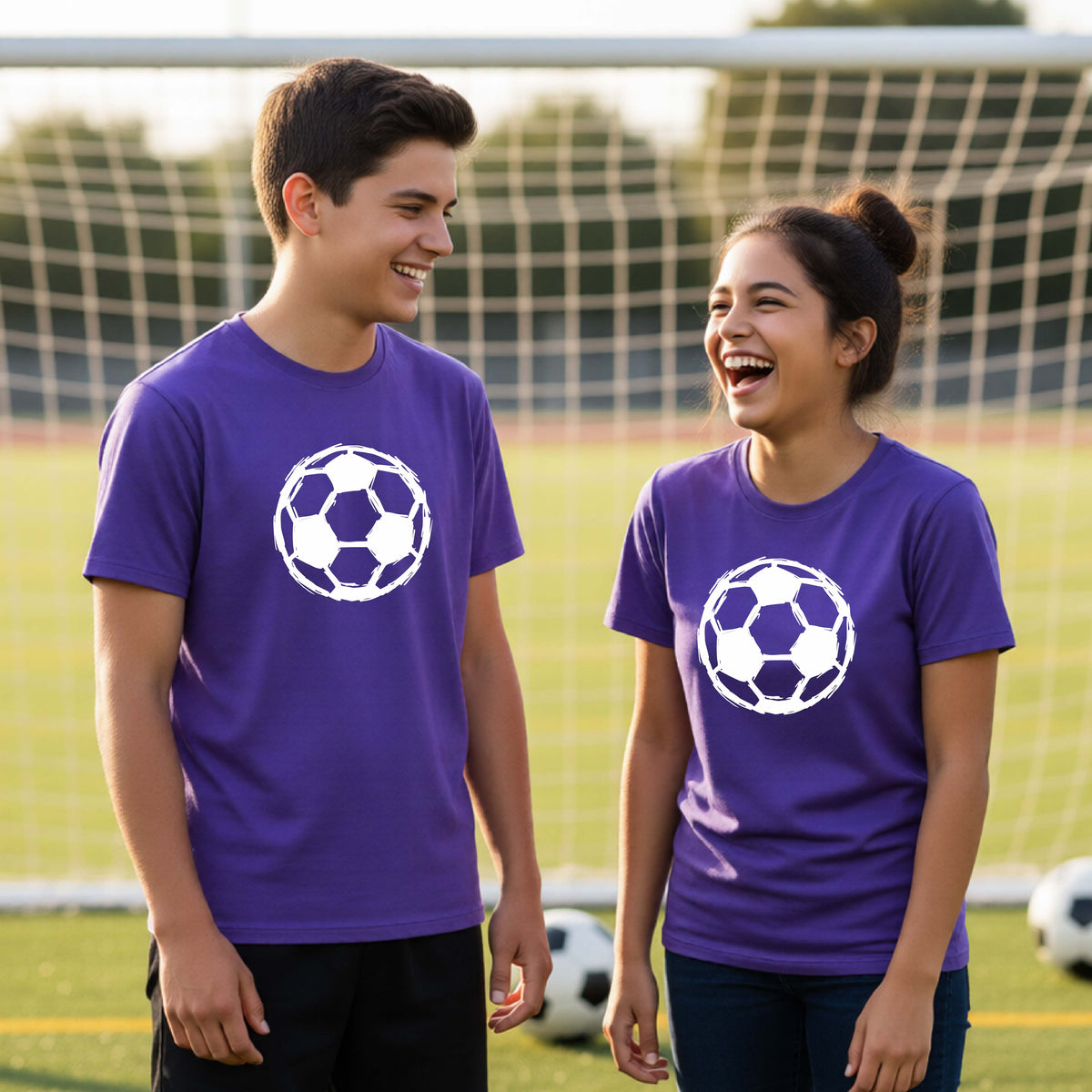 Two smiling young athletes standing on a soccer field wearing matching purple t-shirts with a white soccer ball graphic on the chest.