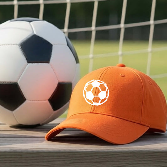 An orange baseball cap with a white soccer ball graphic on the front, resting on a wooden surface in front of a soccer ball and net on an outdoor field.