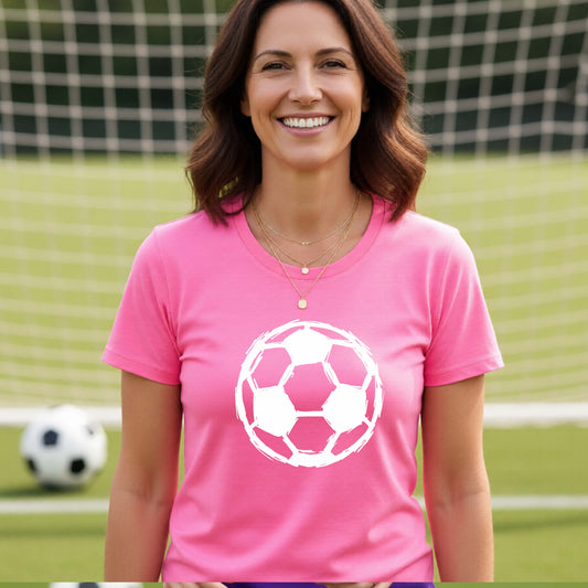 A smiling woman wearing a pink short-sleeve t-shirt with a white soccer ball srtistic graphic on the chest, standing on a soccer field with a goal net and ball in the background.