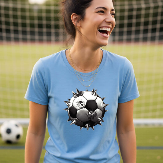 A smiling woman wearing a blue short-sleeve t-shirt standing on a soccer field in front of a goal net with a white and black graphic of a soccer ball with extreme spikes coming out, printed on the front.