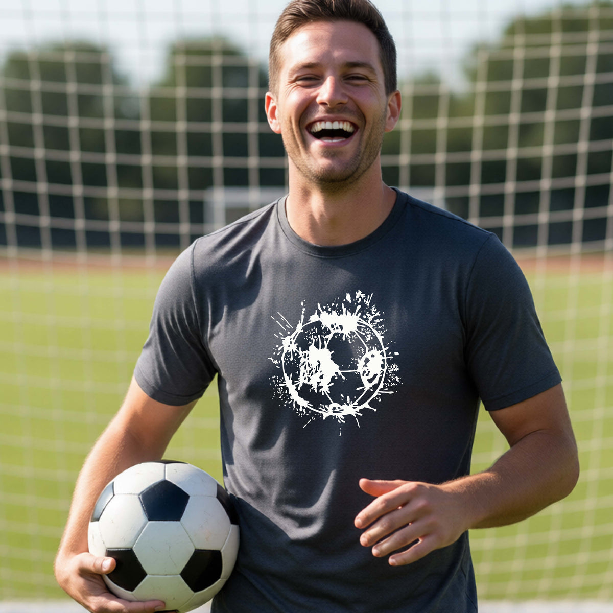 A man wearing a dark gray short-sleeve t-shirt with a white artistic splatter looking soccer ball graphic, standing in front of a soccer goal with a soccer ball in the background.