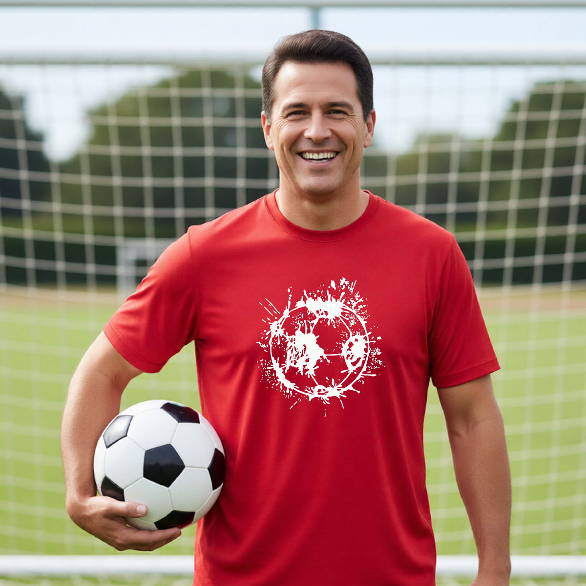 A man wearing a red short-sleeve t-shirt with a white artistic splatter looking soccer ball graphic, standing in front of a soccer goal with a soccer ball in the background.