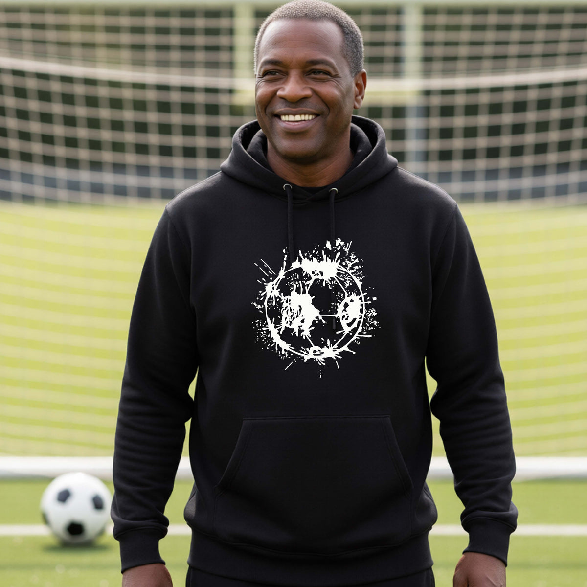 A man wearing a black hoodie pullover with a white artistic splatter looking soccer ball graphic, standing in front of a soccer goal with a soccer ball in the background. 