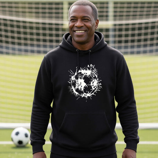A man wearing a black hoodie pullover with a white artistic splatter looking soccer ball graphic, standing in front of a soccer goal with a soccer ball in the background. 