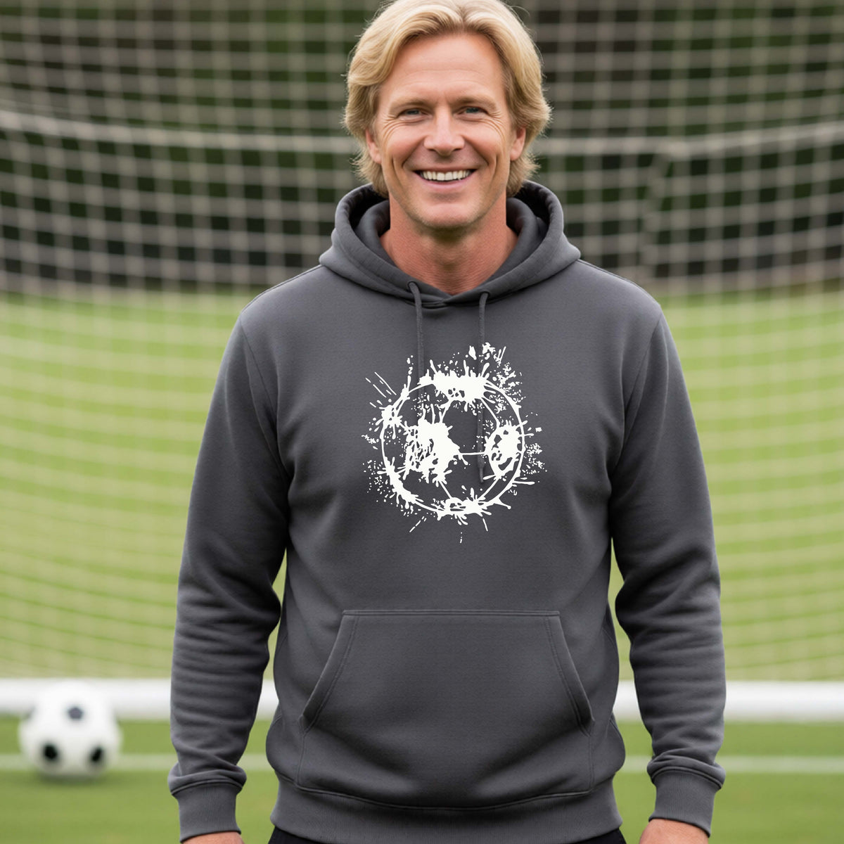 A man wearing a dark gray hoodie pullover with a white artistic splatter looking soccer ball graphic, standing in front of a soccer goal with a soccer ball in the background. 