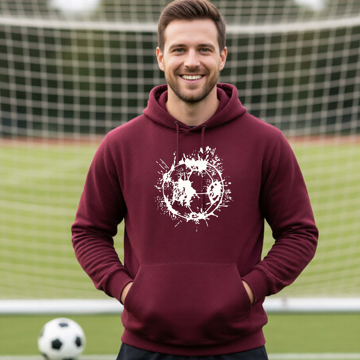 A man wearing a maroon hoodie pullover with a white artistic splatter looking soccer ball graphic, standing in front of a soccer goal with a soccer ball in the background.
