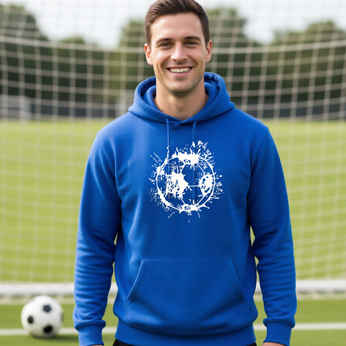 A man wearing a royal blue hoodie pullover with a white artistic splatter looking soccer ball graphic, standing in front of a soccer goal with a soccer ball in the background. 