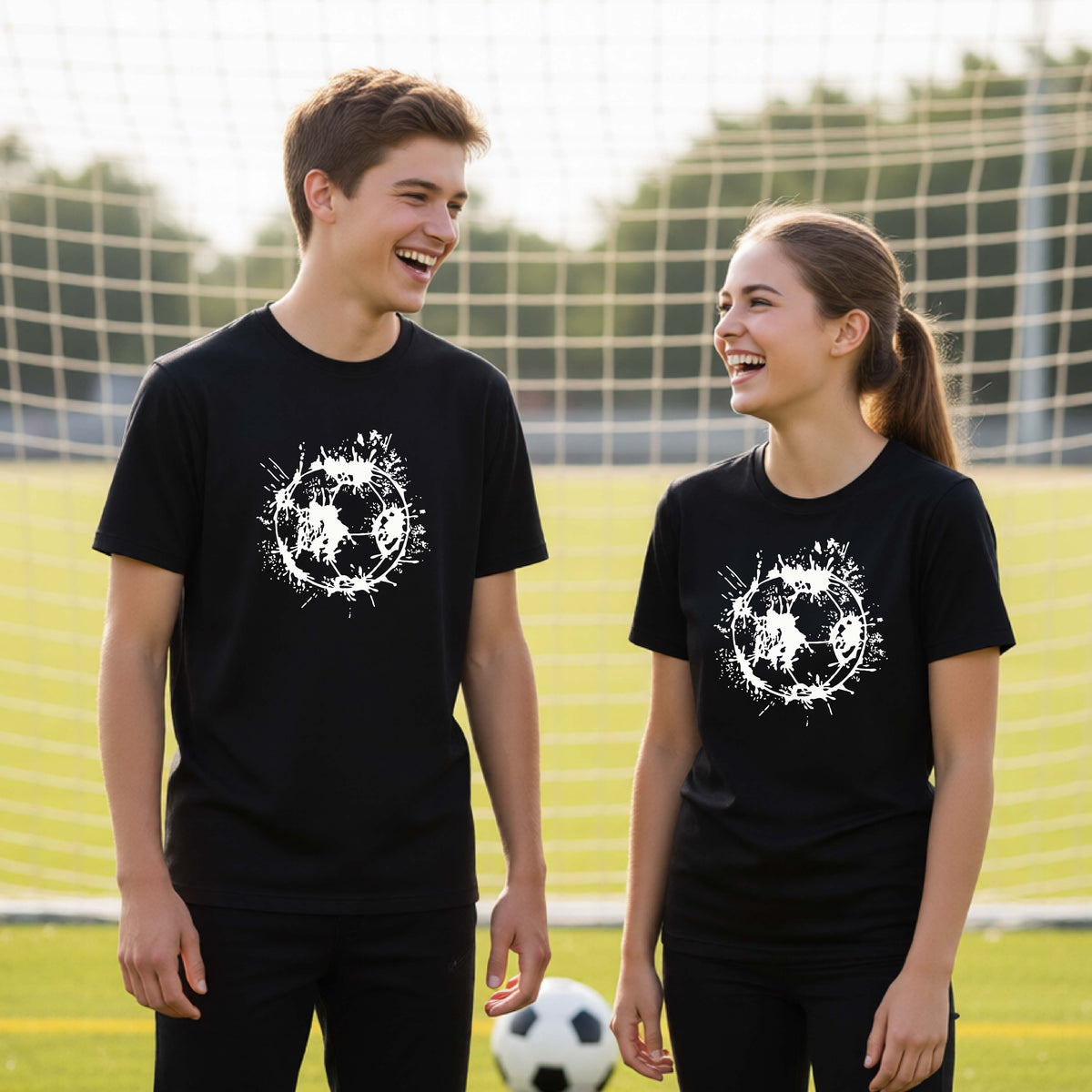 Two young athletes wearing matching black short-sleeve t-shirts with a white artistic splatter looking soccer ball graphic, standing in front of a soccer goal with a soccer ball in the background.