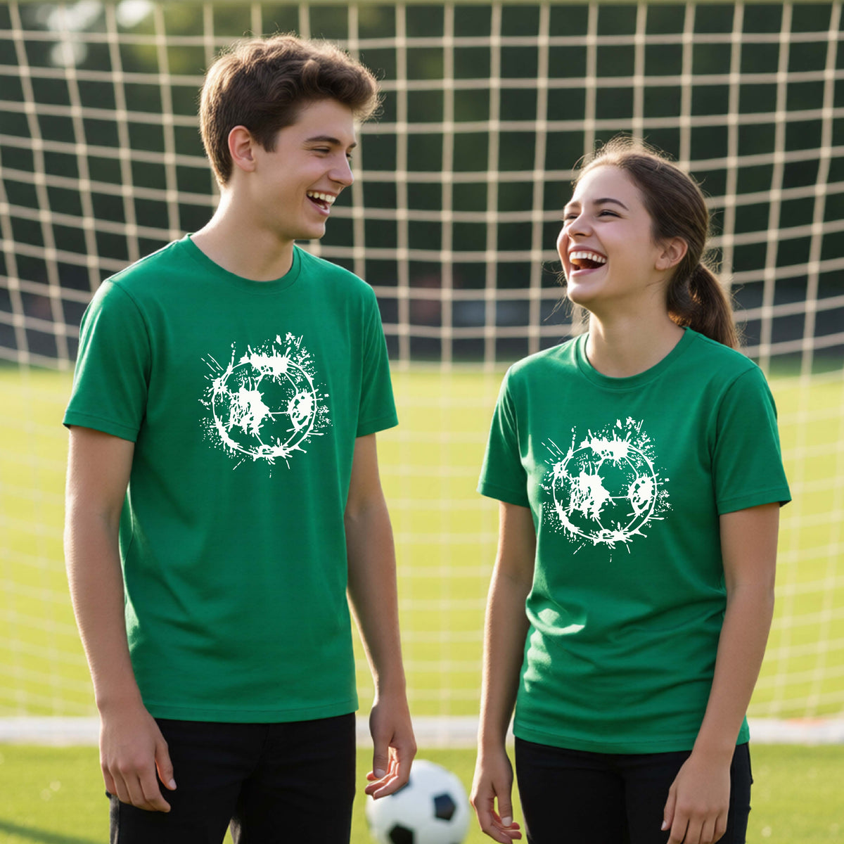 Two young athletes wearing matching green short-sleeve t-shirts with a white artistic splatter looking soccer ball graphic, standing in front of a soccer goal with a soccer ball in the background.