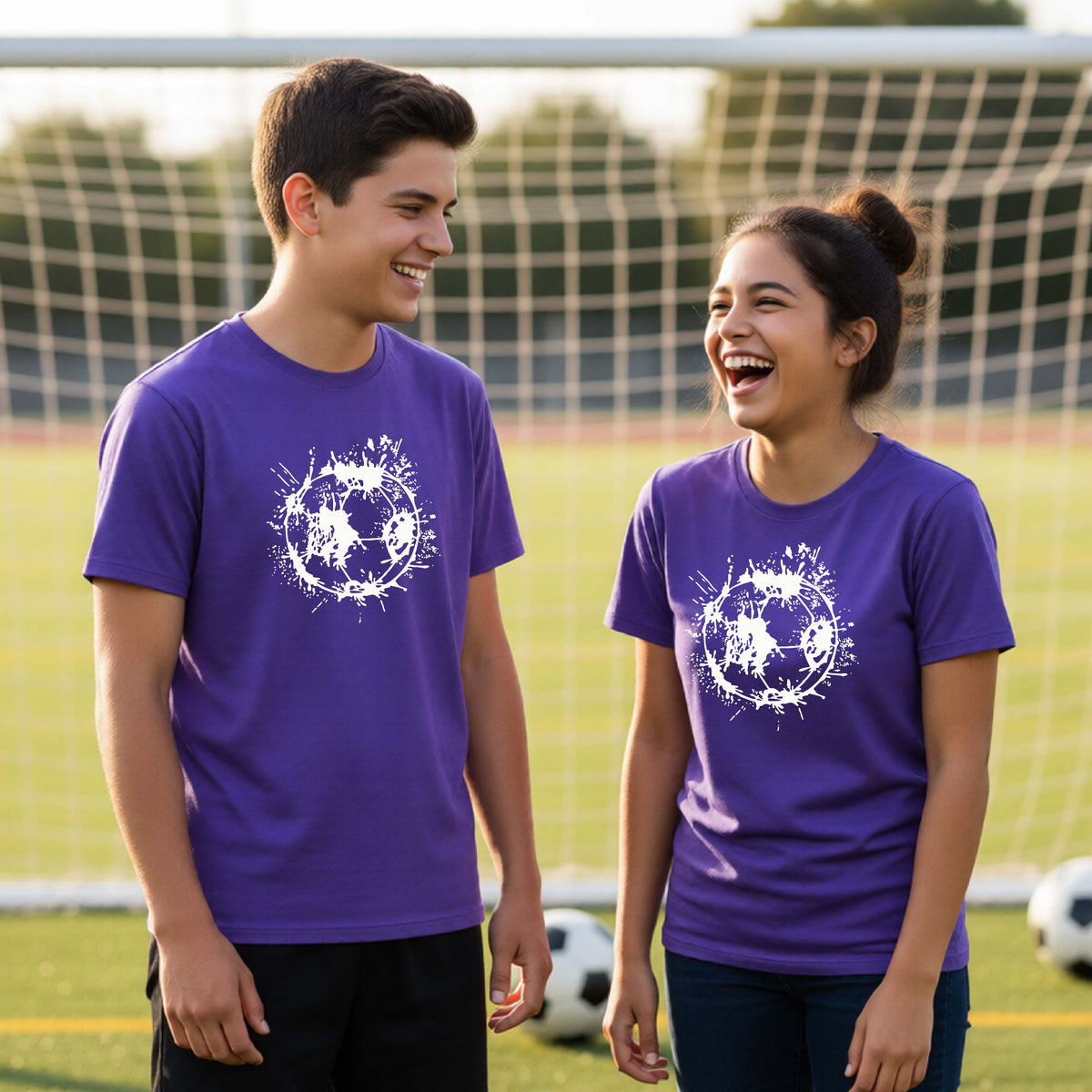 Two young athletes wearing matching purple short-sleeve t-shirts with a white artistic splatter looking soccer ball graphic, standing in front of a soccer goal with a soccer ball in the background.