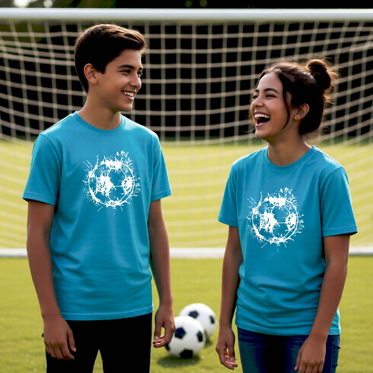 Two young athletes wearing matching bright blue short-sleeve t-shirts with a white artistic splatter looking soccer ball graphic, standing in front of a soccer goal with a soccer ball in the background.