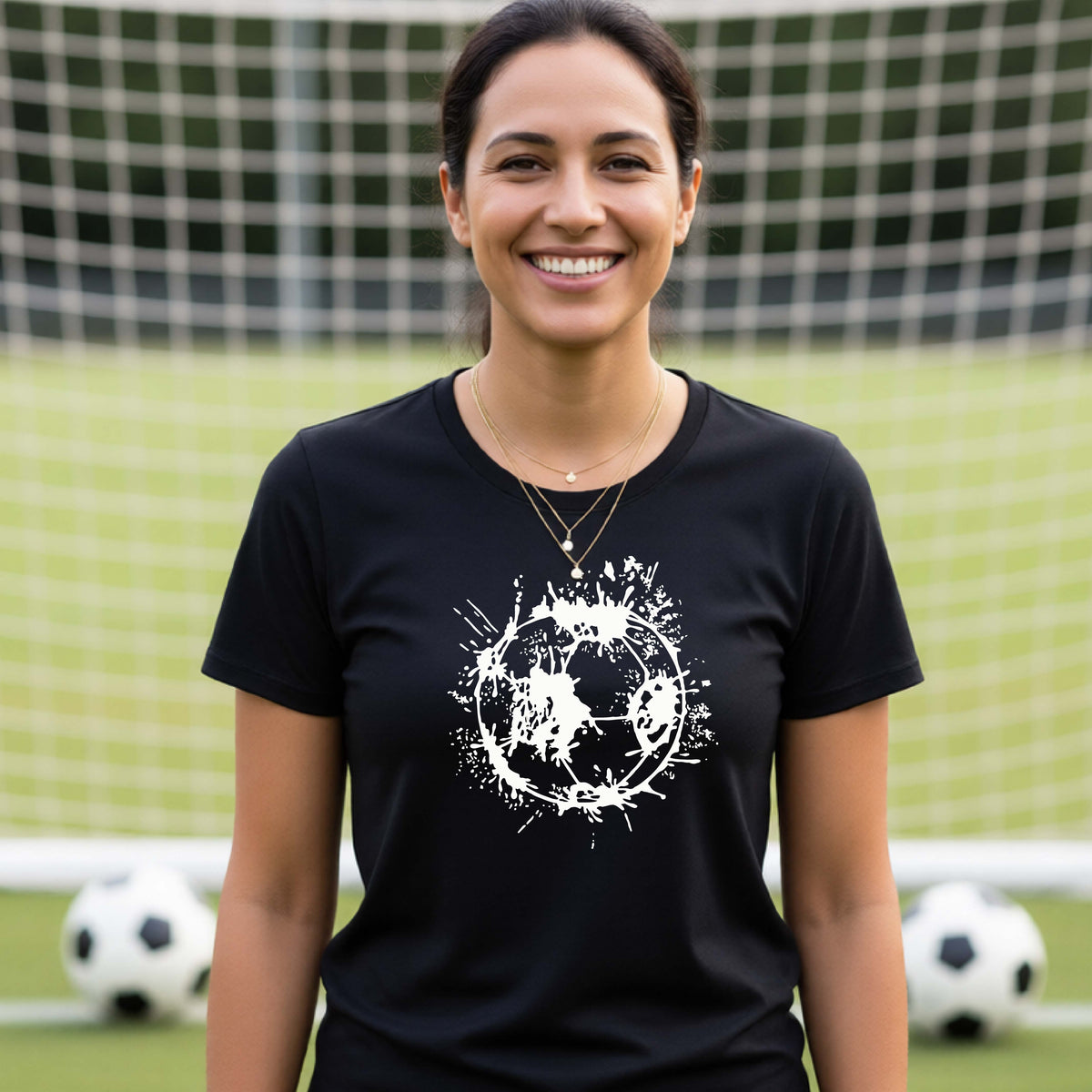 A woman wearing a black short-sleeve t-shirt with a white artistic splatter looking soccer ball graphic, standing in front of a soccer goal with a soccer ball in the background.