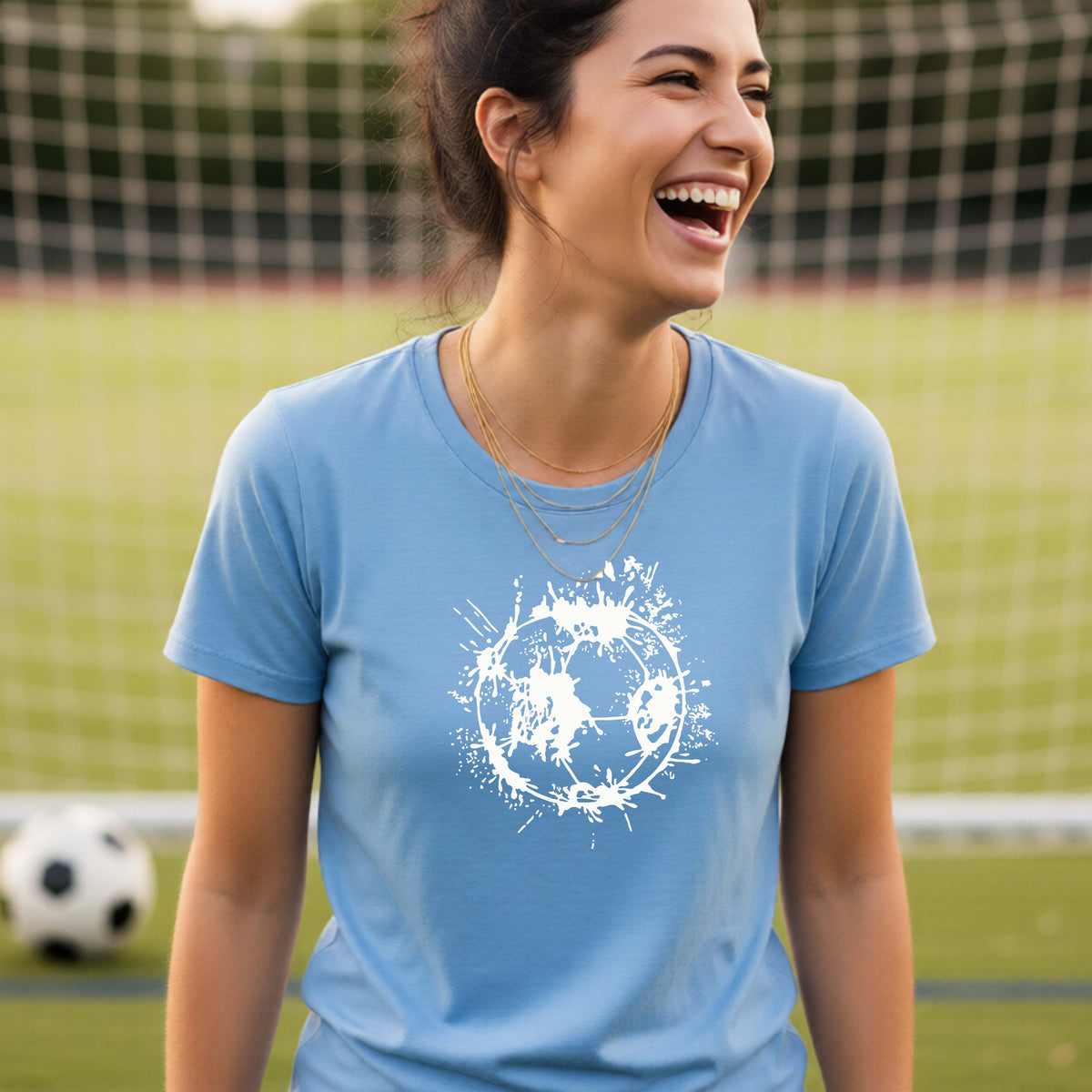 A woman wearing a blue short-sleeve t-shirt with a white artistic splatter looking soccer ball graphic, standing in front of a soccer goal with a soccer ball in the background.