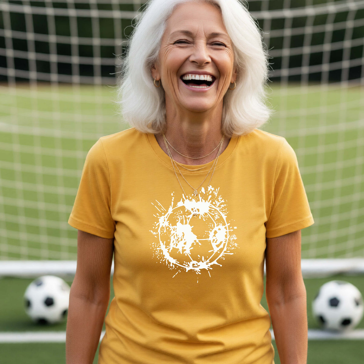 A woman wearing a yellow gold short-sleeve t-shirt with a white artistic splatter looking soccer ball graphic, standing in front of a soccer goal with a soccer ball in the background.