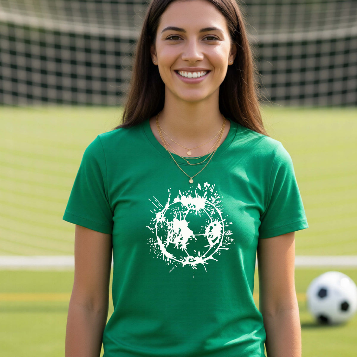 A woman wearing a green short-sleeve t-shirt with a white artistic splatter looking soccer ball graphic, standing in front of a soccer goal with a soccer ball in the background.