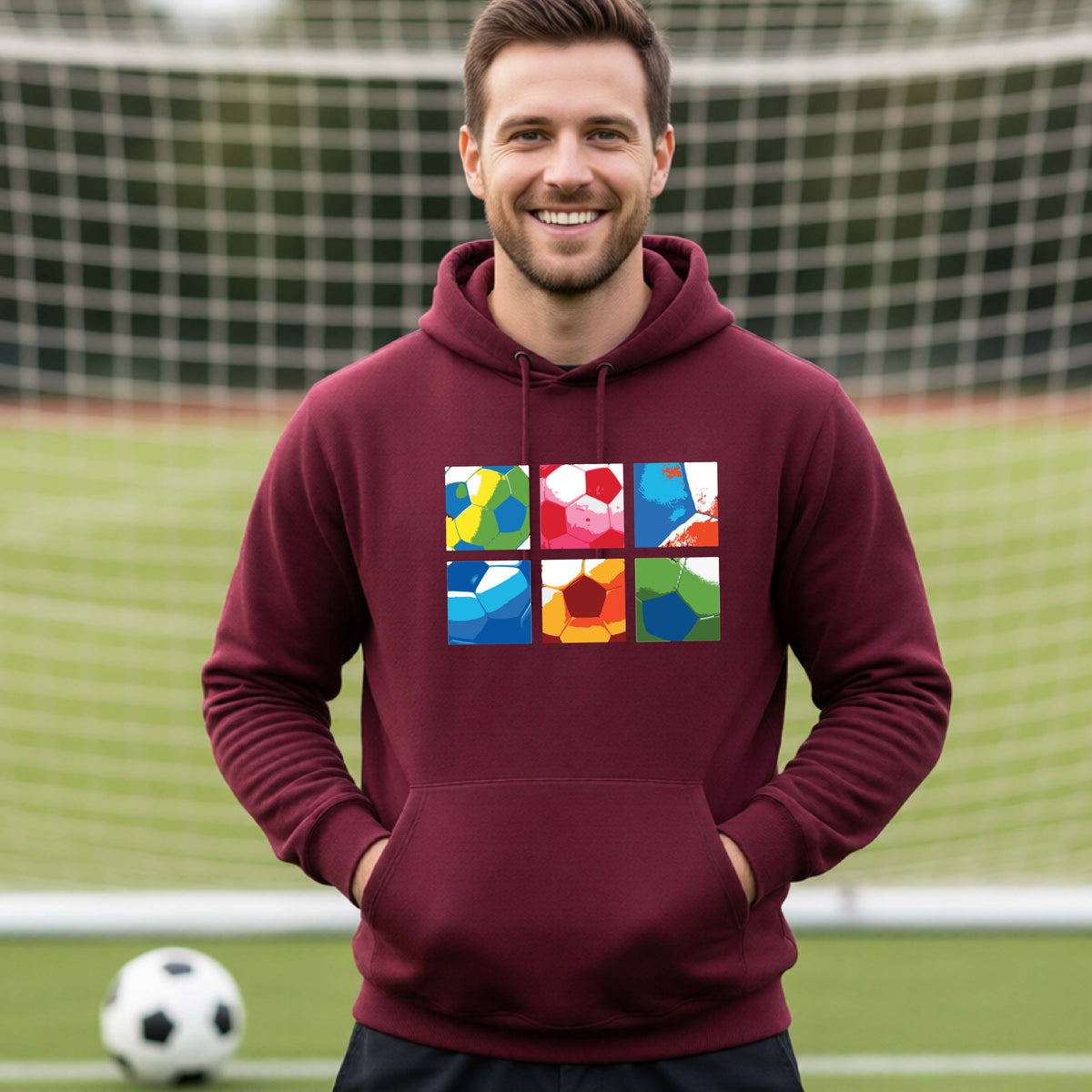 A smiling man wearing a maroon hoodie pullover standing on a soccer field in front of a goal net. with a colorful graphic of a 2x3 arrangement of squares of soccer balls, printed on the chest.