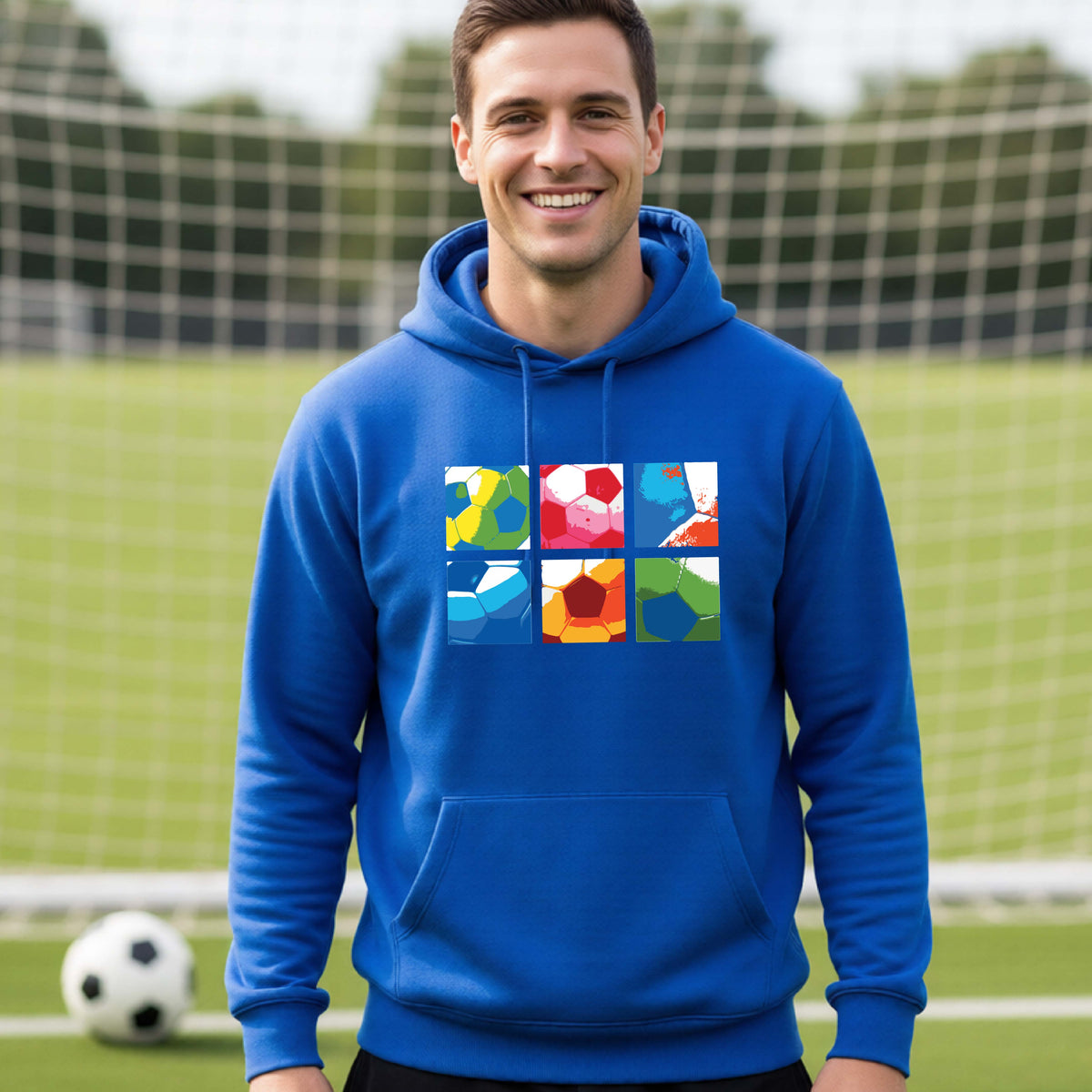 A smiling man wearing a royal blue hoodie pullover standing on a soccer field in front of a goal net. with a colorful graphic of a 2x3 arrangement of squares of soccer balls, printed on the chest.