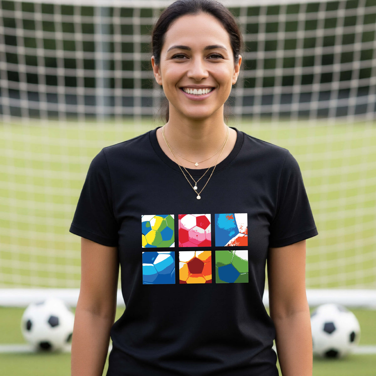 A smiling woman wearing a black short-sleeve t-shirt standing on a soccer field in front of a goal net. with a colorful graphic of a 2x3 arrangement of squares of soccer balls, printed on the chest.