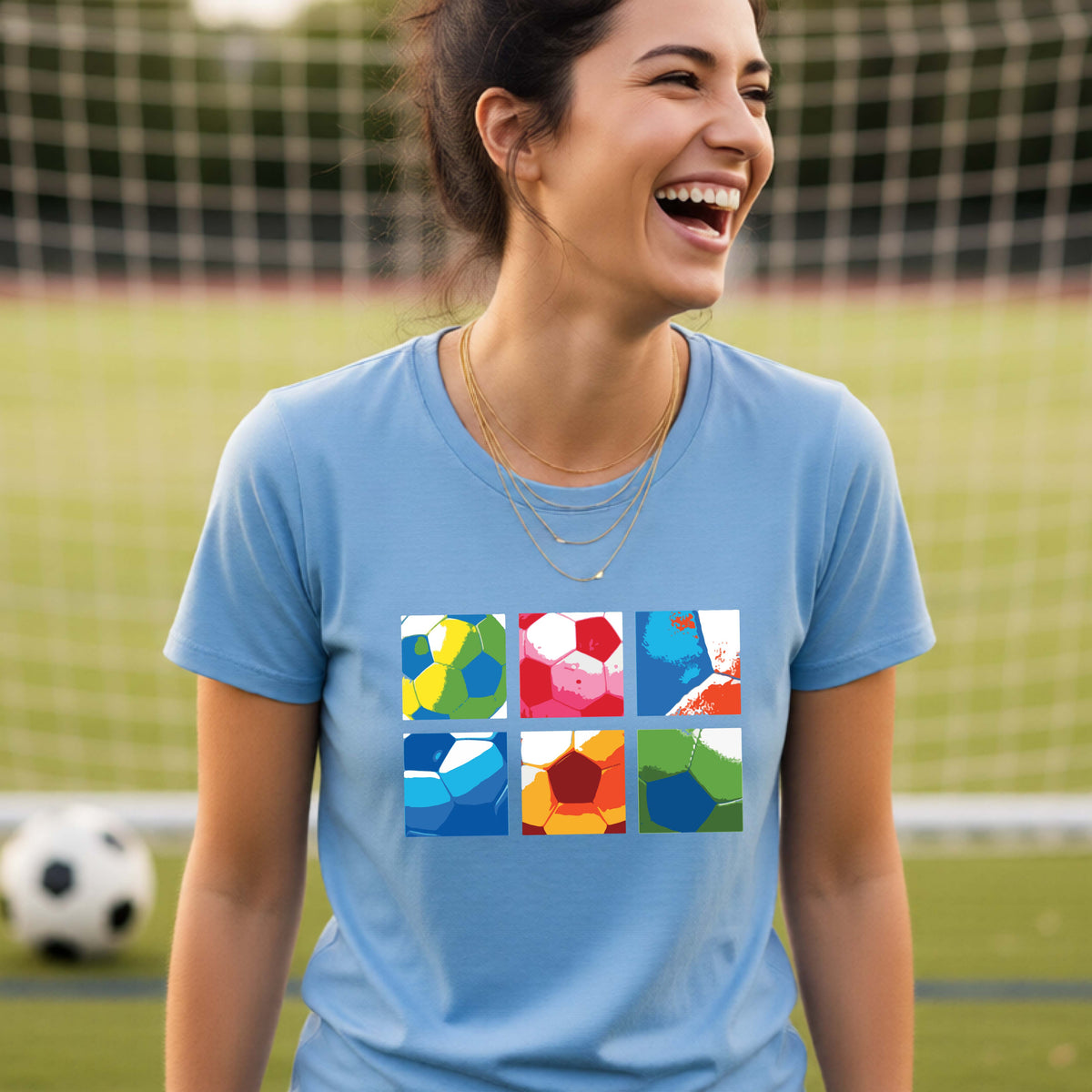 A smiling woman wearing a blue short-sleeve t-shirt standing on a soccer field in front of a goal net. with a colorful graphic of a 2x3 arrangement of squares of soccer balls, printed on the chest.