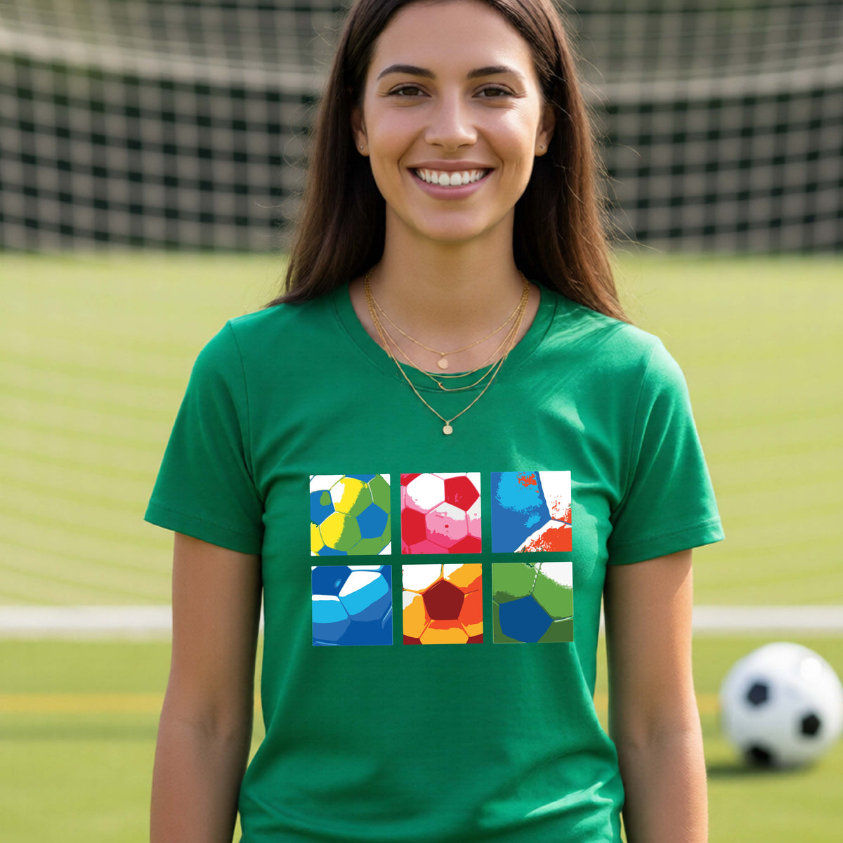 A smiling woman wearing a green short-sleeve t-shirt standing on a soccer field in front of a goal net. with a colorful graphic of a 2x3 arrangement of squares of soccer balls, printed on the chest.