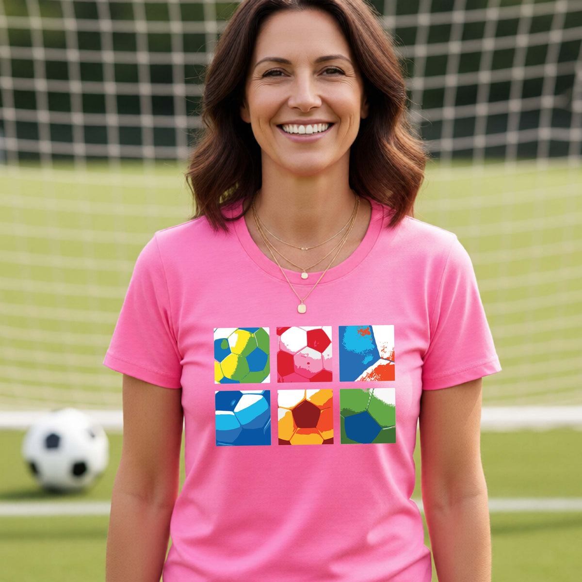 A smiling woman wearing a pink short-sleeve t-shirt standing on a soccer field in front of a goal net. with a colorful graphic of a 2x3 arrangement of squares of soccer balls, printed on the chest.
